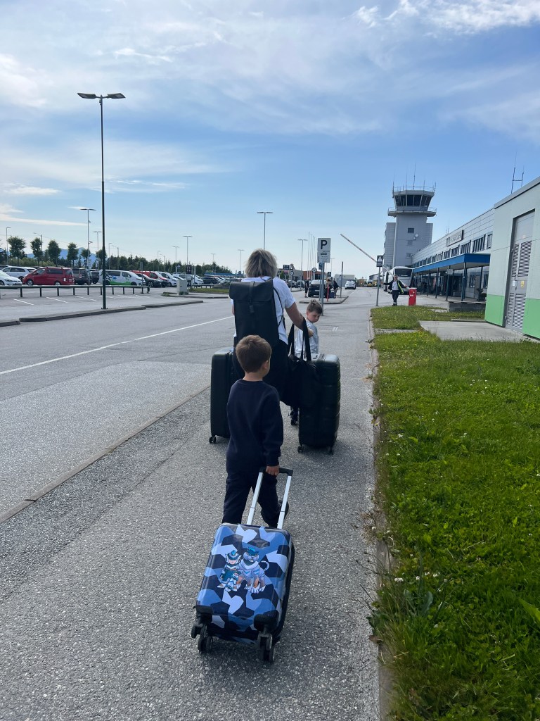 A family walking towards an airport with luggage, including a child pulling a cartoon-themed suitcase, under a clear sky.
