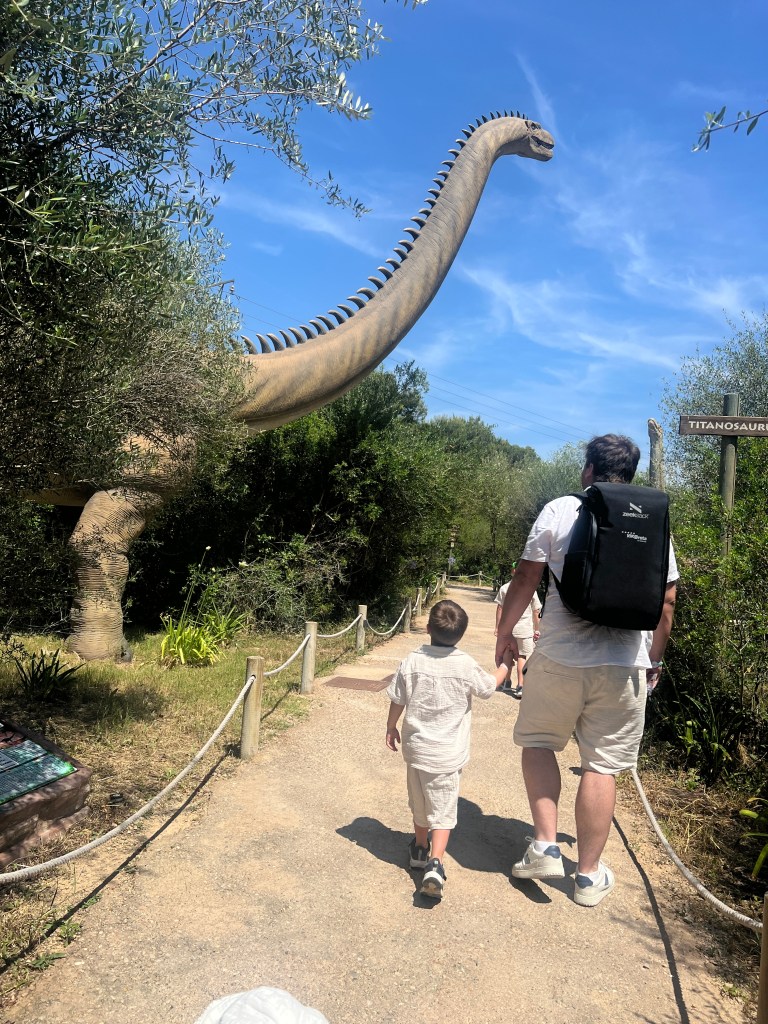 A parent and child walking hand in hand on a path, with a large dinosaur statue in the background surrounded by greenery.