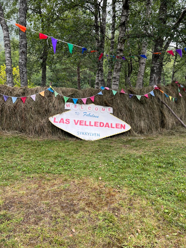 A welcome sign for 'Las Velledalen' in a green, grassy area adorned with colorful bunting and surrounded by trees.