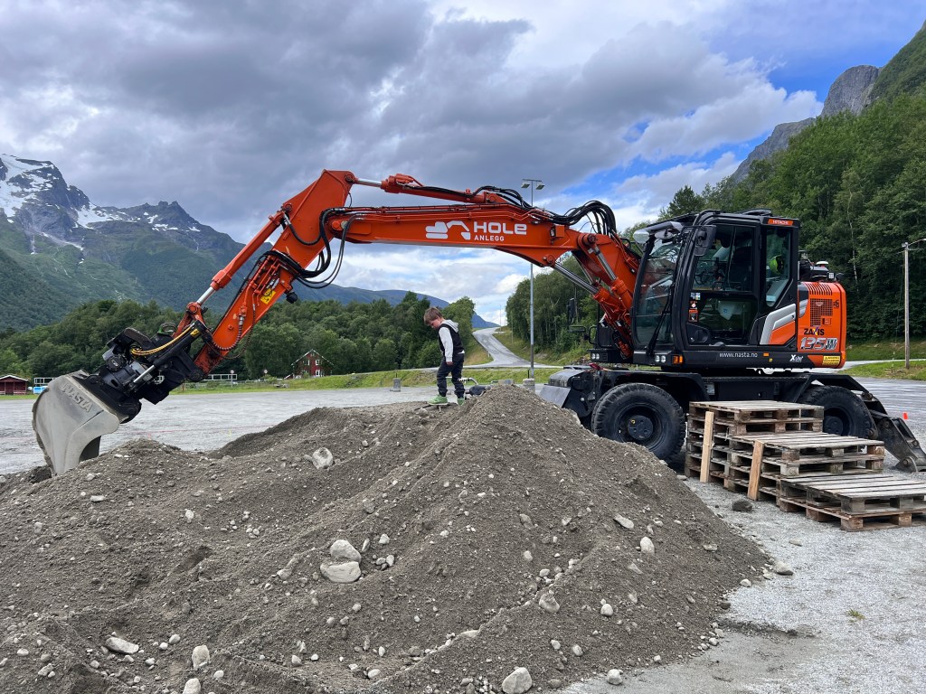 A construction site featuring an excavator with a large bucket lifting dirt, and a child playing near a pile of gravel.