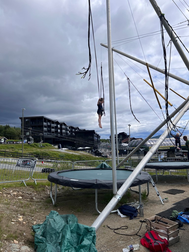 Child hanging in a harness from a bungee trampoline frame with a trampoline beneath, in a recreational area with cloudy skies and buildings in the background.