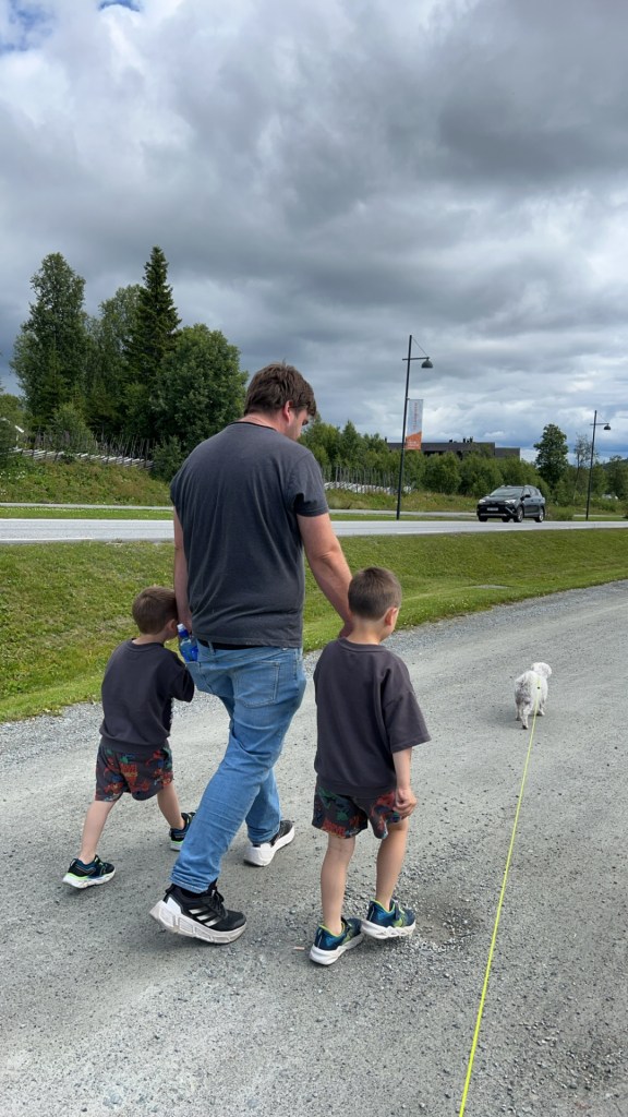 A man walking with two young boys and a small dog on a gravel path, surrounded by greenery and cloudy skies.