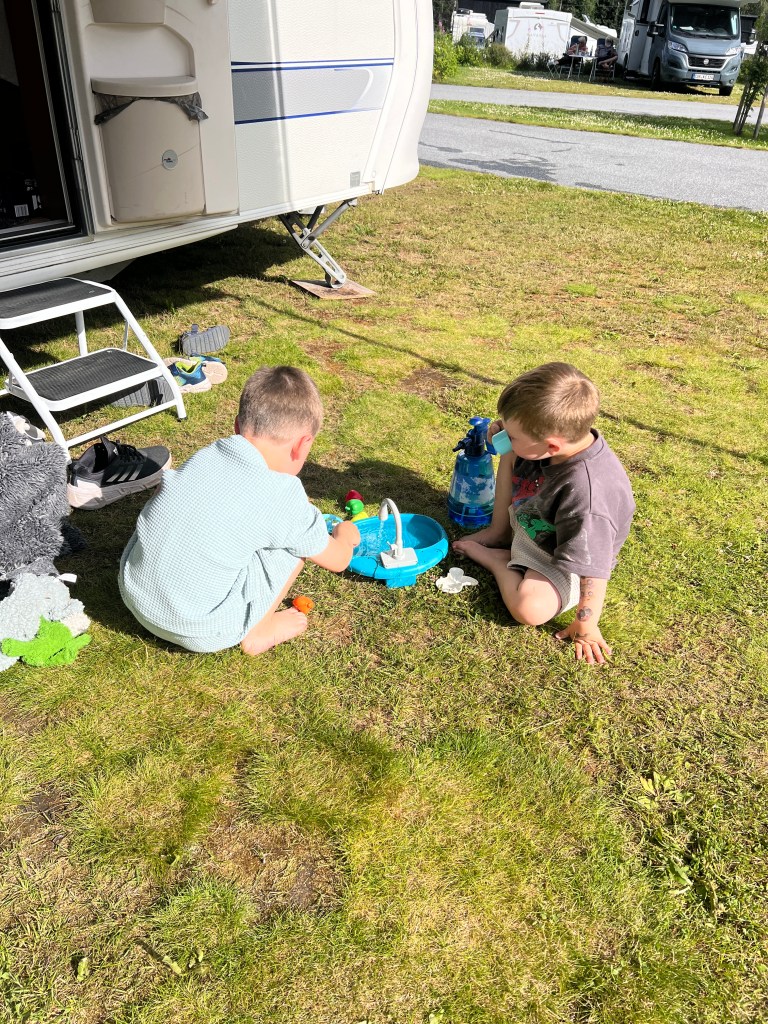 Two young children playing with water toys on grass outside a caravan.
