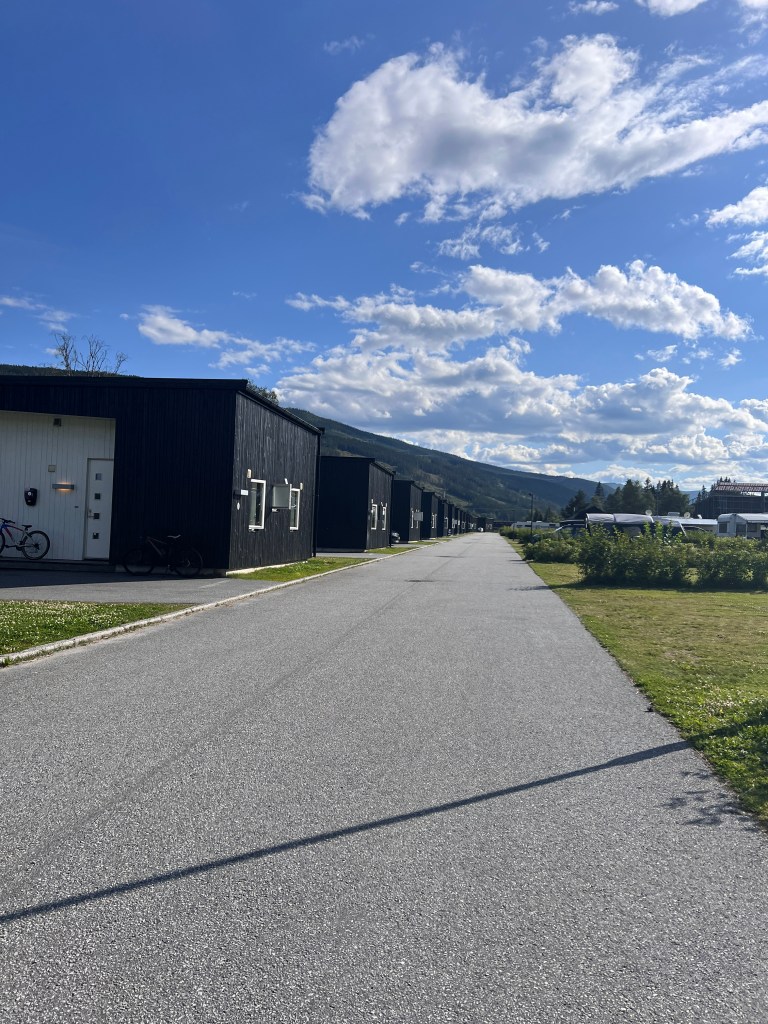 A scenic view of a quiet road lined with black cabins under a blue sky with fluffy clouds and mountains in the background.