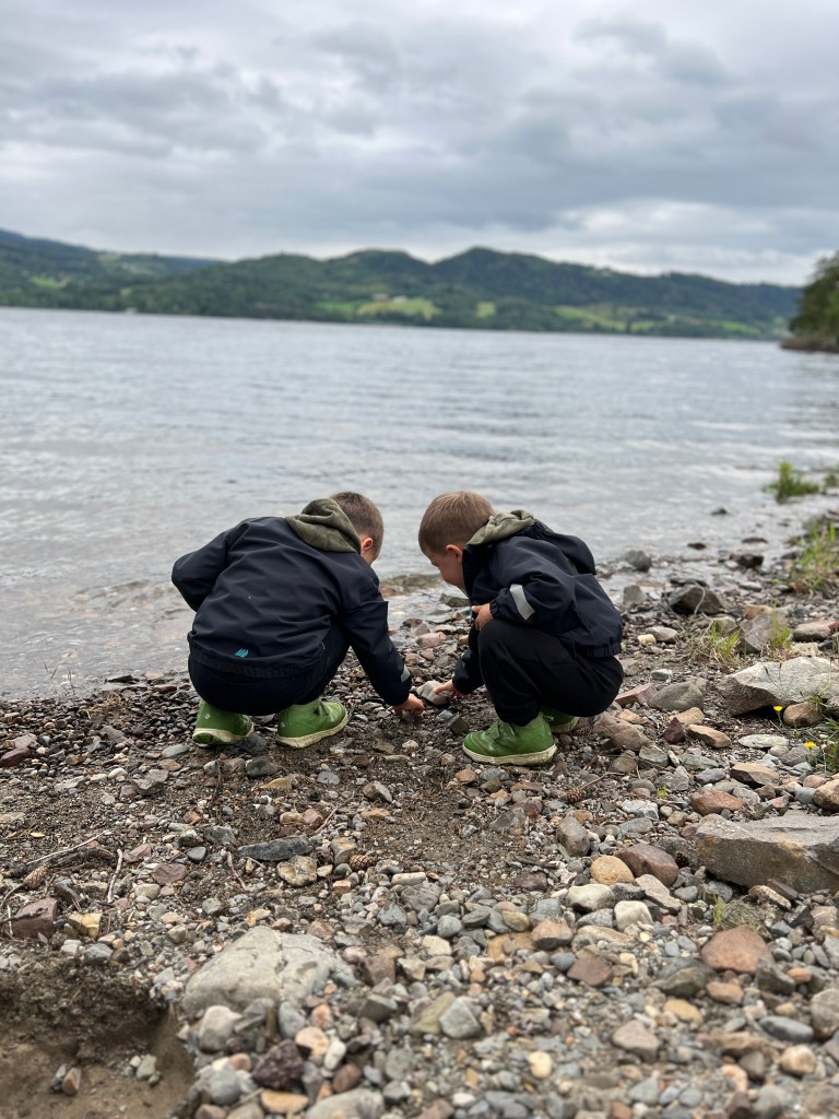 Two children exploring the rocky shoreline near a lake, wearing matching dark outfits and green boots.