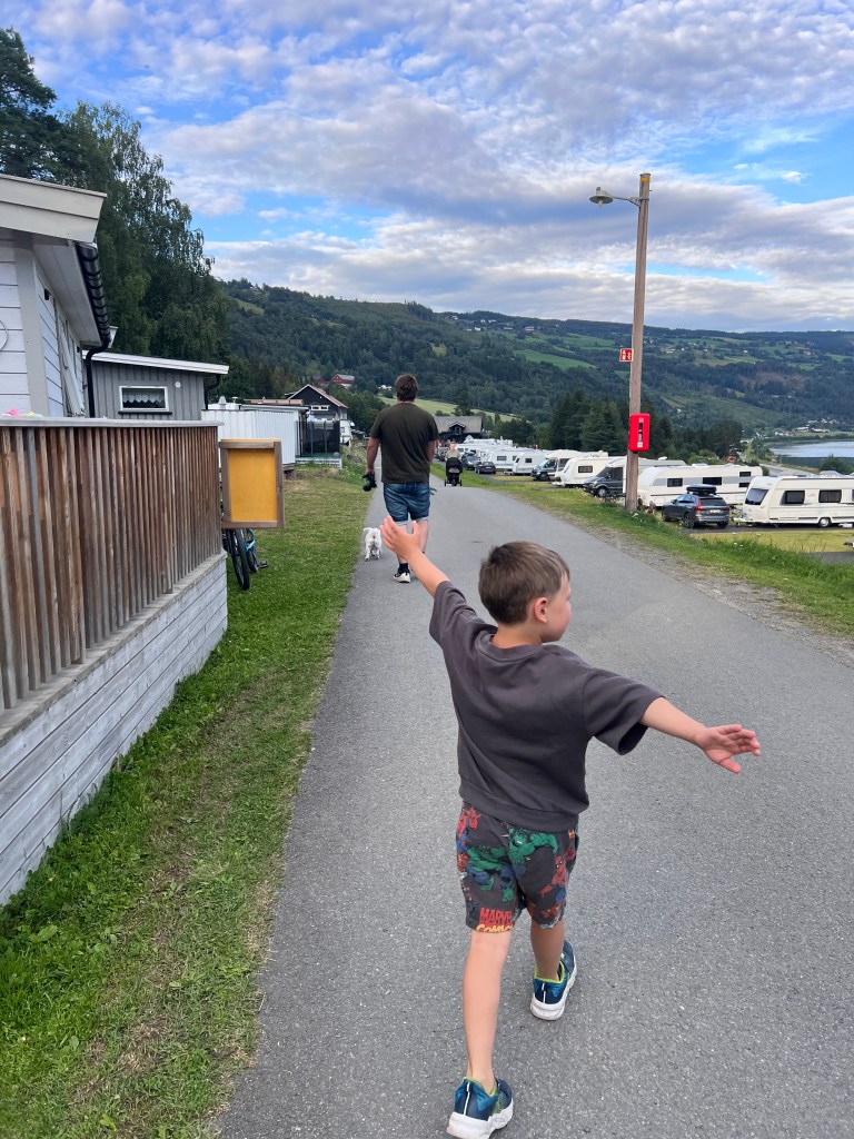A young boy joyfully walks down a path, reaching out his arm, while a man follows behind him in a campsite setting surrounded by green hills and cloudy skies.